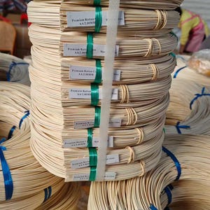 Op de afbeelding: Close-up van gebundeld, lichtgekleurd rotan, vastgebonden met groene en blauwe banden. Elk pakket is voorzien van de tekst "Premium Rattan AA/2.00MM". Het rotan is een natuurlijk materiaal, waarschijnlijk voor knutselen of meubels.