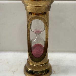 May include: A brass hourglass featuring a polished metallic frame with a cylindrical design. The central glass bulb contains fine pink sand, with a small amount remaining in the top chamber and the majority collected in the bottom chamber.