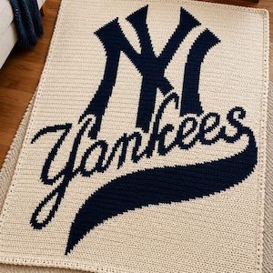 Könnte beinhalten: Eine rechteckige Häkeldecke mit cremefarbenem Hintergrund, auf der das dunkelmarineblaue Logo der New York Yankees und das Wort Yankees in Schreibschrift in der Mitte abgebildet sind. Das Textil hat eine sichtbare Maschenstruktur und einen einfachen Rand.