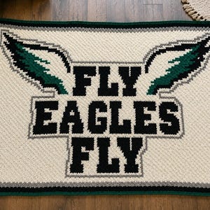 May include: A rectangular crochet blanket featuring a cream-coloured background with a dark green and black eagle wing graphic at the top. The text FLY EAGLES FLY is centred in bold black block letters, all bordered by a dark green and grey trim.