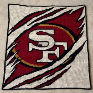 May include: A square crochet blanket featuring a red, gold, and white San Francisco 49ers logo centered on a cream background. The design includes a claw-mark effect with black and red accents, finished with a solid black border around the edge.