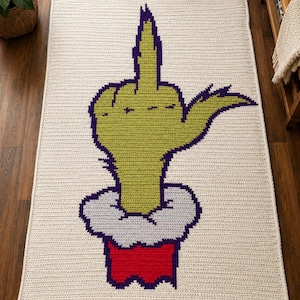 May include: A rectangular crochet blanket with an off-white background featuring a large, centered graphic of a lime green hand with purple outlines making an obscene gesture. The hand emerges from a white fur-like cuff and a red sleeve base.
