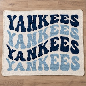 Könnte beinhalten: Eine rechteckige Häkeldecke mit cremefarbenem Hintergrund und dem Wort YANKEES, das viermal in einem wellenförmigen, gestapelten Muster wiederholt wird. Der Text wechselt zwischen dunkelmarineblauem und hellblauem Garn für ein strukturiertes Design.
