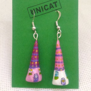 Puede incluir: Un par de pendientes hechos a mano con un diseño de casa de ensueño. Las casas están pintadas en tonos de rosa, morado y blanco con detalles dorados. Los pendientes están unidos a ganchos plateados.