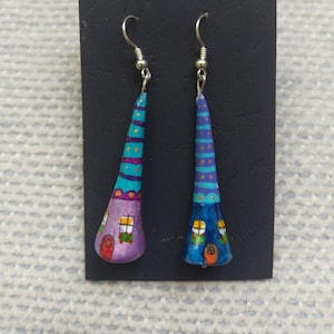 Puede incluir: Un par de pendientes hechos a mano con diseños de casas caprichosas. Los pendientes están pintados en colores brillantes, incluyendo morado, azul, turquesa y amarillo. Cada casa tiene una pequeña ventana y una puerta.