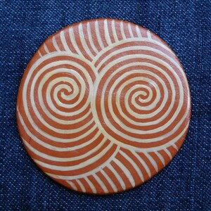May include: A round, terracotta-colored coaster with a white spiral design. The coaster features two overlapping spirals, creating a unique and intricate pattern.