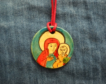 Puede incluir: Un colgante redondo de cerámica con una imagen pintada de una mujer y un niño. La mujer lleva una túnica roja y azul y el niño lleva una túnica amarilla. El colgante está en un cordón rojo.