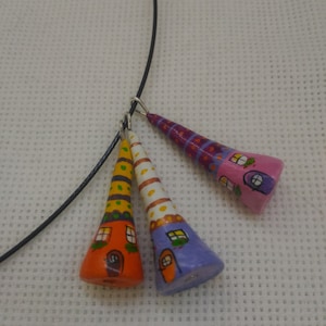May include: A whimsical necklace featuring three hand-painted, cone-shaped pendants resembling colorful houses. The pendants are in orange, yellow, and purple, each with unique window and door details. The necklace has a black cord.