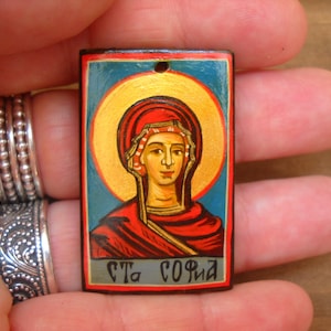 Può includere: Un'icona in legno dipinta a mano di una donna con un'aureola, vestita con una tunica rossa e dorata. L'icona ha uno sfondo blu e il testo "Cта София" è scritto in basso.