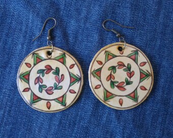May include: A pair of round earrings with a cream-colored background and a decorative design. The earrings feature a central wreath of red and green leaves, surrounded by a starburst pattern with red and green triangles. The earrings hang from silver-colored hooks.