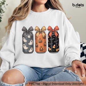 Könnte beinhalten: Weißes Sweatshirt mit drei Halloween-Soda-Dosen. Die Dosen sind grau, orange und schwarz, jede mit einer Schleife und gefüllt mit gruseligen Grafiken wie Geistern, Kürbissen, Fledermäusen und Zuckermais. Das b.dots STUDIO-Logo befindet sich oben rechts.