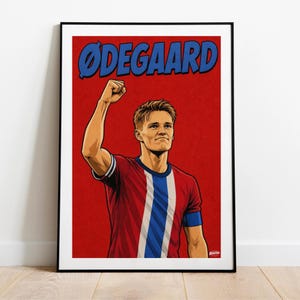 May include: A framed poster featuring a stylized illustration of a soccer player with a raised fist. The player wears a red jersey with a blue and white vertical stripe. The text "ØDEGAARD" is printed in large blue letters against a red background.