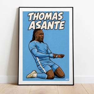 May include: A framed poster featuring a cartoon illustration of Thomas Asante in a light blue soccer uniform. The player is kneeling on a light blue background, with the name "THOMAS ASANTE" in bold white letters above him.