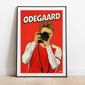May include: A framed art print featuring a stylized illustration of a person holding a camera. The person is wearing a red and white shirt with gold accents. The word "ODEGAARD" is printed in white on a red background.