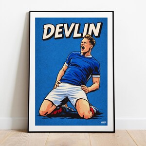 Op de afbeelding: Een ingelijste poster met een gestileerde illustratie van een voetballer in een blauw shirt en witte shorts, die viert. Het woord "DEVLIN" staat in vette, witte letters met een zwarte omlijning tegen een blauwe achtergrond.