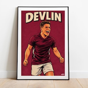 Op de afbeelding: Een ingelijste kunstprint met een gestileerde illustratie van een voetballer in een bordeauxrood shirt met witte accenten en een witte short. De speler viert met opgeheven armen, met de naam "DEVLIN" erboven.