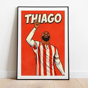 May include: A framed art print with a comic book style illustration of a footballer in a red and white striped shirt, pointing upwards. The name "THIAGO" is printed in bold white letters above. The background is a solid red colour.
