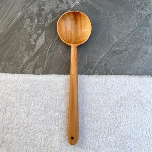 May include: A wooden cooking spoon with a round bowl and a long handle. The spoon is made of light brown wood and has a hole at the end of the handle. The spoon is designed for stirring and serving food.