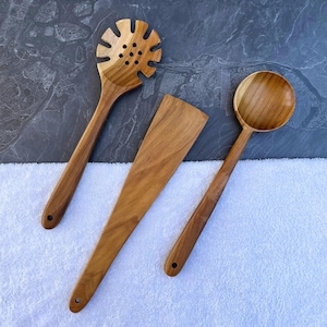Può includere: Tre utensili da cucina in legno: un cucchiaio forato, una spatola e un mestolo. Gli utensili sono in legno marrone chiaro e hanno manici lunghi. Il cucchiaio forato ha un disegno a forma di fiore sulla testa. La spatola è piatta e rettangolare. Il mestolo ha una ciotola rotonda.