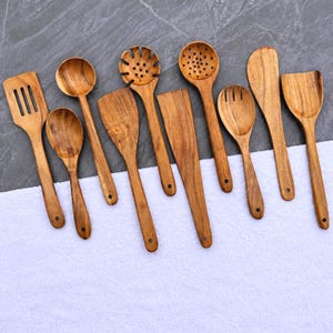Könnte beinhalten: Ein Set aus neun Küchenutensilien aus Holz, bestehend aus verschiedenen Löffeln, Pfannenwendern, einem Schlitzwender und einem Nudellöffel. Jedes Teil hat eine natürliche Holzmaserung und ein kleines Loch am Griffende zum Aufhängen.
