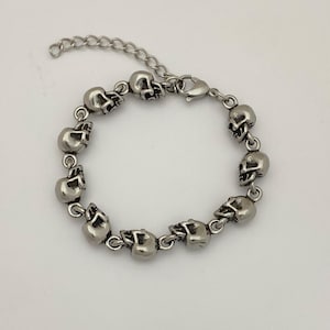 May include: Silver-toned bracelet featuring a series of small skull charms. The bracelet has a chain extension for adjustable sizing and a lobster clasp closure. The skulls are detailed with eye sockets, noses, and teeth.