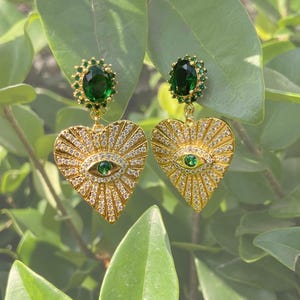 May include: Gold-toned heart-shaped earrings with a central eye design. The earrings feature a green gemstone eye and are adorned with small, sparkling crystals. The top of the earrings have a green gemstone surrounded by smaller green stones.
