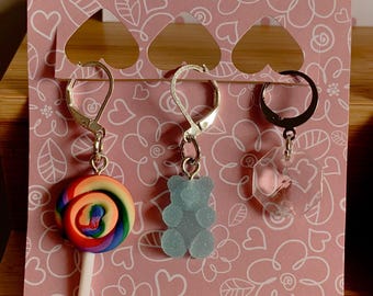 Può includere: Tre paia di orecchini pendenti su una carta rosa con motivi a cuore e fiori. Un orecchino presenta un lecca-lecca colorato, un altro un orsetto gommoso blu e il terzo una forma geometrica trasparente. Gli orecchini hanno ganci color argento.