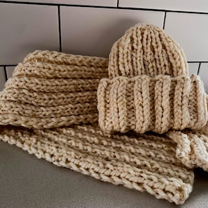 May include: A chunky knit cream-coloured beanie hat and a matching long scarf. Both items are crafted from thick yarn with a ribbed texture and feature small, embedded gold-coloured metallic flecks throughout the fabric for a subtle shimmer.