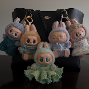 Puede incluir: Cinco llaveros de peluche con forma de conejito en varios colores, incluyendo azul, beige y lavanda, con trajes de ganchillo a juego. Cada llavero tiene una expresión única y está unido a un bolso tote de cuero negro.