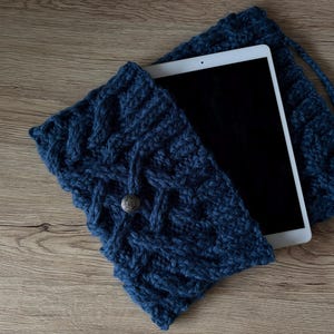 Peut inclure: Une housse de tablette en tricot de câble bleu foncé avec une pochette assortie à cordon de serrage. La housse a une fermeture à bouton décoratif. Une tablette blanche est partiellement visible à l'intérieur. Sur une surface en bois.