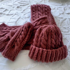 Puede incluir: Un conjunto de punto a juego en color rosa empolvado, que incluye gorro, bufanda y guantes sin dedos. El conjunto presenta un diseño de punto trenzado y se muestra sobre una superficie blanca texturizada.