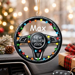 May include: A circular hanging ornament shaped like a black steering wheel decorated with colorful Christmas lights. The center features the text NEW DRIVER 2026, and the top section displays the name Max in white lettering against a clear background.