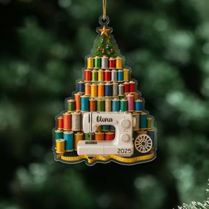 May include: A Christmas tree ornament featuring a sewing machine and spools of colorful thread. The ornament is shaped like a Christmas tree, with a star on top. The name "Olivia" and the year "2025" are visible on the ornament.