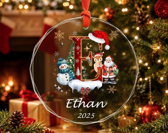 May include: A round, clear acrylic Christmas ornament featuring a large letter E decorated with a Santa hat, a snowman, a reindeer, and Santa Claus. The ornament includes the name Ethan and the year 2025 printed in white, suspended by a red ribbon.
