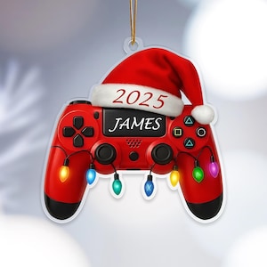 May include: Red video game controller Christmas ornament with a Santa hat and string lights. The ornament has the name "JAMES" and the year "2025" printed on it. The controller has black accents and colorful lights.