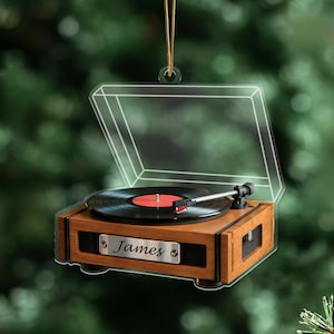 Könnte beinhalten: Ein Weihnachtsornament in Form eines Plattenspielers in Holzoptik mit einer transparenten Kunststoffabdeckung. Der Plattenspieler hat eine schwarze Schallplatte mit roter Mitte. Der Name "James" steht auf einem silbernen Namensschild. Ein goldener Faden ist zum Aufhängen befestigt.