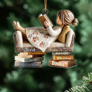 May include: Ornament of a girl reading a book while reclining on a couch. The ornament is supported by a stack of books. The text "Olivia" and "Just a Girl Who Loves Books" are visible.