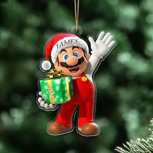 May include: A Christmas tree decoration featuring a cartoon character in a Santa hat, red overalls, and white gloves. The ornament holds a green gift box with the year 2025 on it. The name "JAMES" is printed on the hat.
