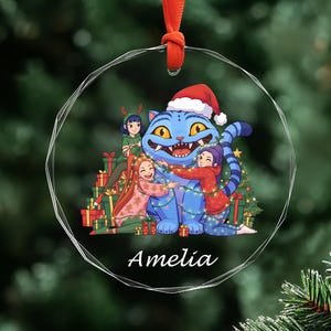 May include: A clear glass Christmas ornament with a red ribbon. The ornament features a blue cartoon cat wearing a Santa hat, surrounded by three people and Christmas presents. The name "Amelia" is written at the bottom.