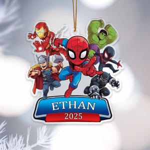 May include: A clear acrylic Christmas ornament featuring a group of cartoon superheroes, including Spider-Man, Iron Man, and Thor. The ornament has a blue banner with the name "ETHAN" and the year "2025". A gold string is attached for hanging.