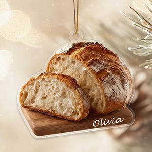 May include: A bread-shaped ornament featuring a loaf of crusty bread and a slice, resting on a wooden cutting board. The ornament has a clear, circular top with a gold string for hanging. The name "Olivia" is written on the cutting board.