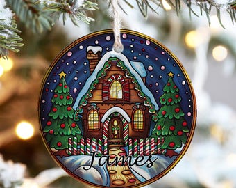 May include: Circular Christmas decoration with a gingerbread house design, snow-covered roof, and trees. The ornament has a blue backdrop with falling snow and the name "James" at the bottom. It is suspended by a twine string.