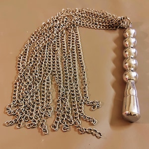 Puede incluir: Borla de cadena de metal plateado con un asa decorativa. La borla se compone de múltiples hebras de cadena, conectadas a una cadena que conduce a un asa con una serie de segmentos redondeados y pulidos. El asa tiene una base cónica.