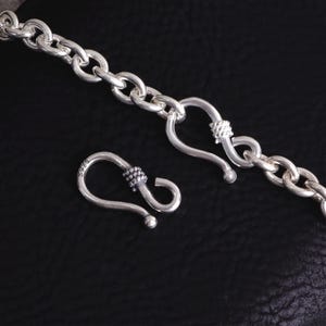 May include: A silver chain necklace with a large clasp. The chain links are oval-shaped and connected. The clasp is a hook and eye design with decorative detailing. The necklace is displayed on a black surface.