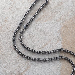 Op de afbeelding: Close-up van een zilverkleurige ketting met een ovaal schakelontwerp. De ketting wordt weergegeven op een getextureerd, lichtgekleurd oppervlak. De ketting heeft een karabijnsluiting.