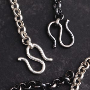 Puede incluir: Primer plano de cierres y eslabones de cadena plateados sobre un fondo oscuro. Los cierres tienen forma de S y las cadenas tienen un diseño de eslabones redondeados. Las joyas parecen ser de metal.