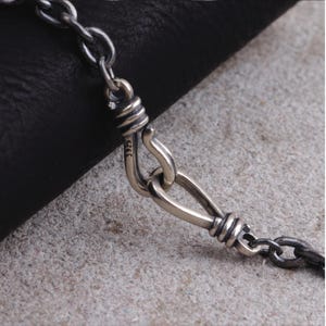 Könnte beinhalten: Nahaufnahme einer silbernen Halskette mit Hakenverschluss. Der Verschluss ist aufwendig mit gewickelten Drahtdetails gestaltet. Kette und Verschluss vor strukturiertem, neutralem Hintergrund.