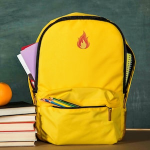 Puede incluir: Una mochila amarilla brillante con cremallera y ribetes negros. La mochila tiene un bolsillo frontal con l&aacute;pices de colores y un peque&ntilde;o dise&ntilde;o de llama bordada. El compartimento principal est&aacute; abierto, revelando cuadernos y un bloc de notas.