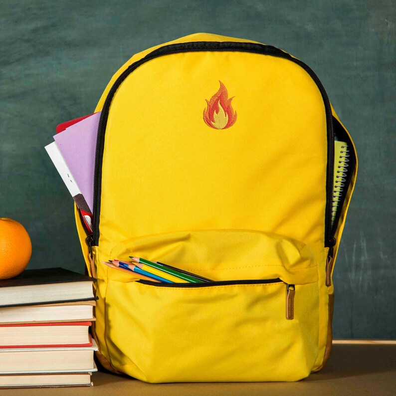 Puede incluir: Una mochila amarilla brillante con cremallera y ribetes negros. La mochila tiene un bolsillo frontal con l&aacute;pices de colores y un peque&ntilde;o dise&ntilde;o de llama bordada. El compartimento principal est&aacute; abierto, revelando cuadernos y un bloc de notas.