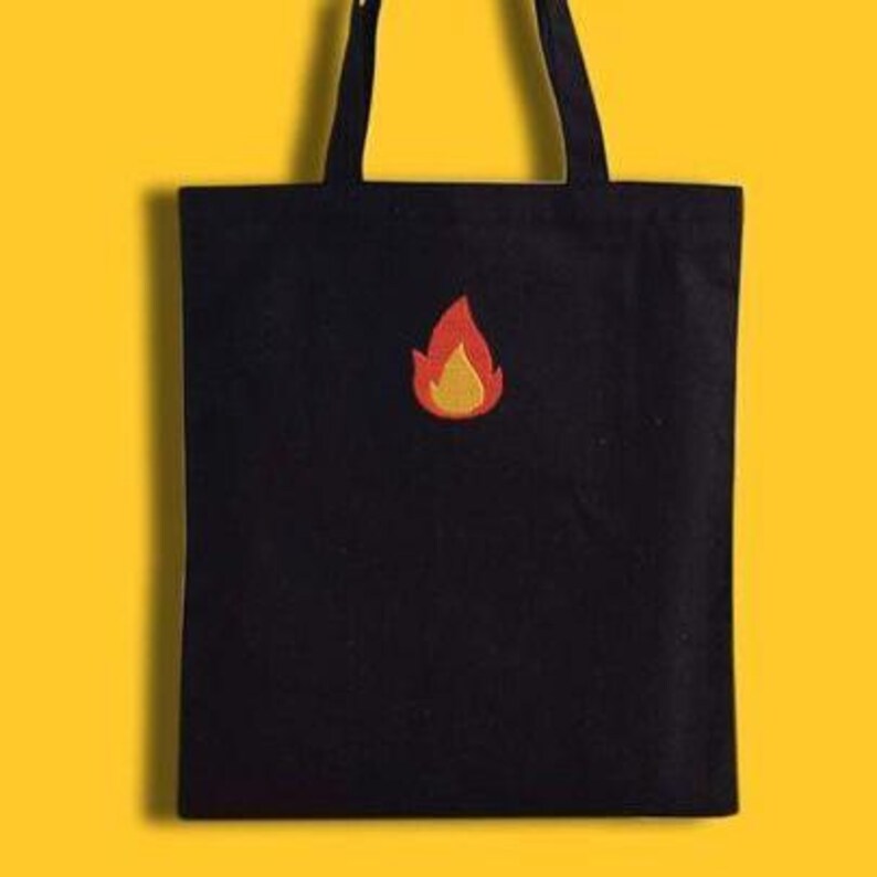Puede incluir: Bolso tote negro con dise&ntilde;o de llama. El bolso tiene una llama roja y amarilla bordada en la parte delantera. Tiene dos asas negras y est&aacute; hecho de un material resistente.